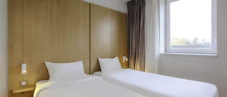 Habitación de hotel con dos camas individuales, cabecera de madera, mesita de noche con lámpara y ventana con cortinas.