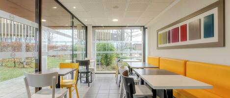 Un comedor moderno con mesas y sillas, un banco amarillo y ventanas amplias con vistas al exterior.