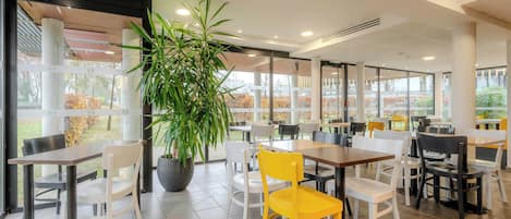 Un espace de restauration intérieur moderne, avec des tables et des chaises, de grandes fenêtres et une plante en pot.