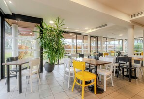 Un espace de restauration intérieur moderne, avec des tables et des chaises, de grandes fenêtres et une plante en pot.