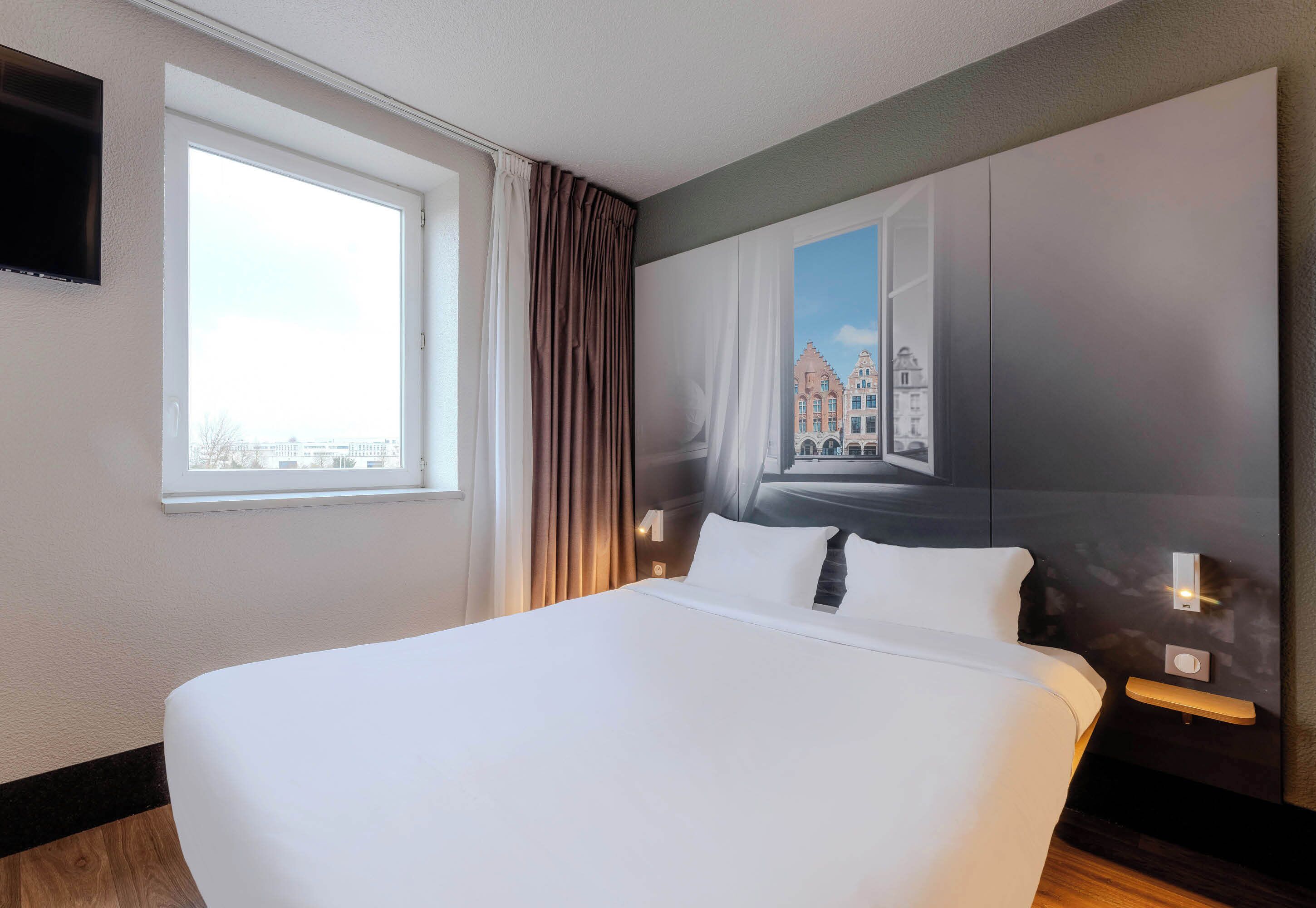 A hotel room with a large bed, a window with curtains, a wall-mounted TV, and a cityscape painting.