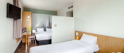 A hotel room with two single beds, a desk with a chair, a flat-screen TV mounted on the wall, and a small refrigerator.