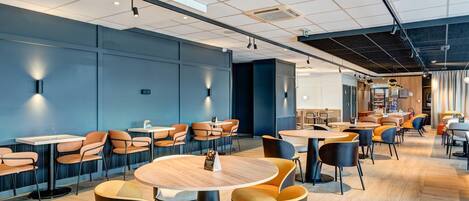 A modern hotel lobby with multiple seating areas, including round tables and chairs, and a visible reception desk.