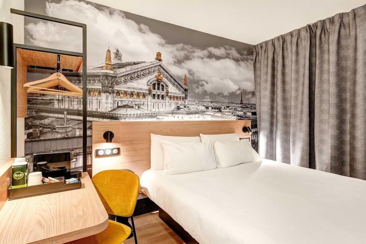 A hotel room with a bed, a desk with a chair, a coffee machine, and a wall mural of a building.