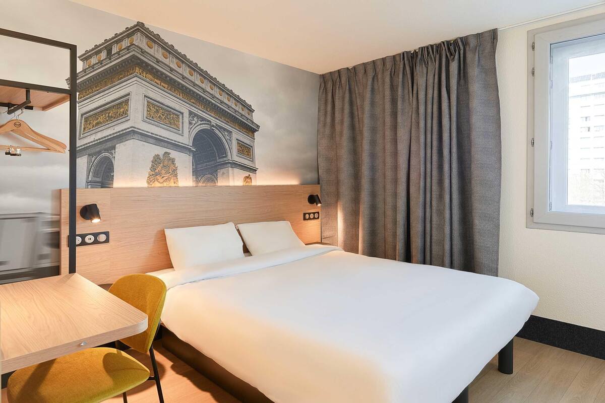 A hotel room with a bed, a desk, a chair, a wardrobe, and a large window.