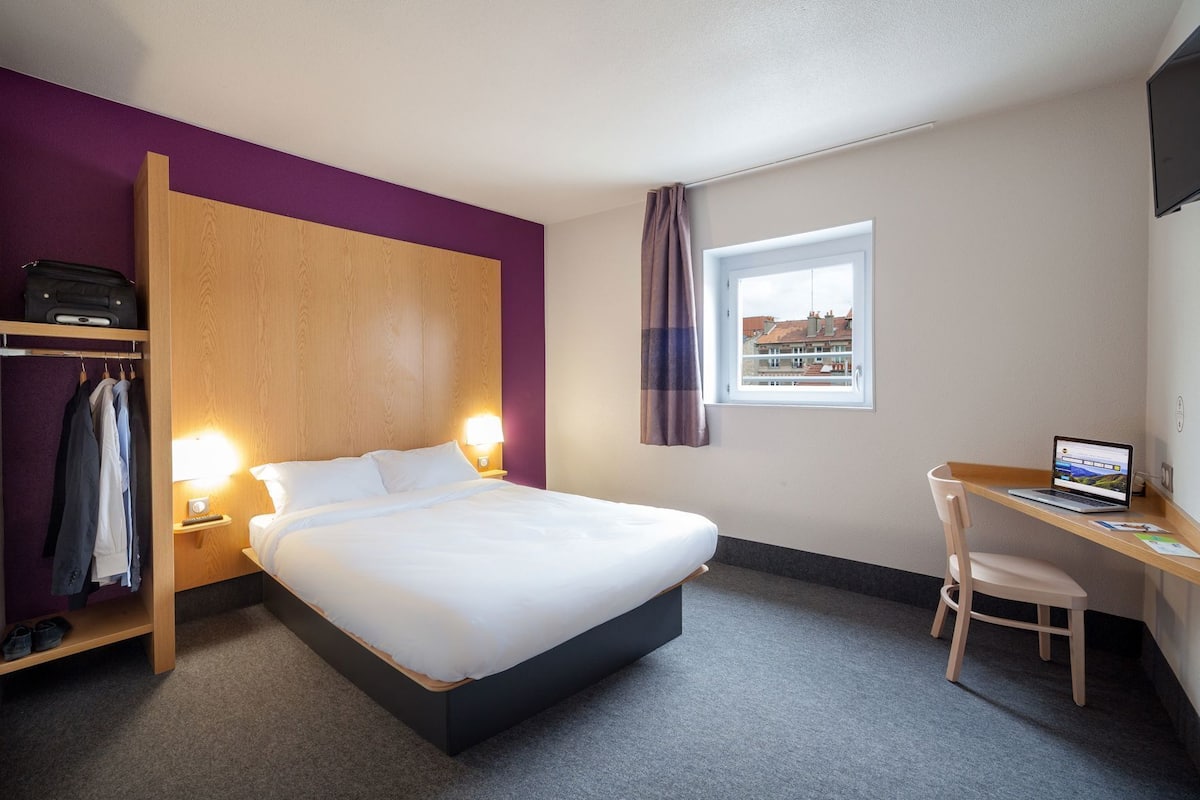 A hotel room with a bed, a desk with a laptop, a wardrobe, and a window with a view.