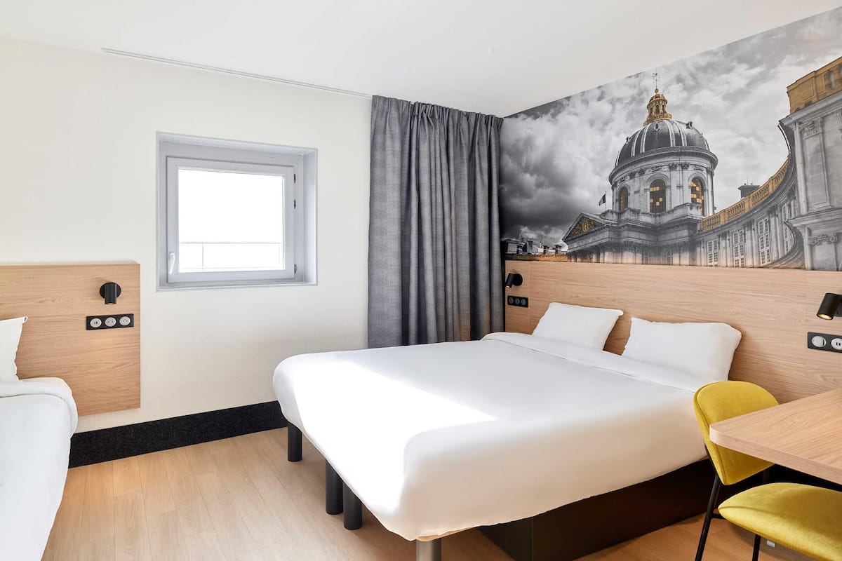 A hotel room with a large bed, a window, a curtain, a wall-mounted lamp, a wooden headboard, a small table, and a yellow chair.