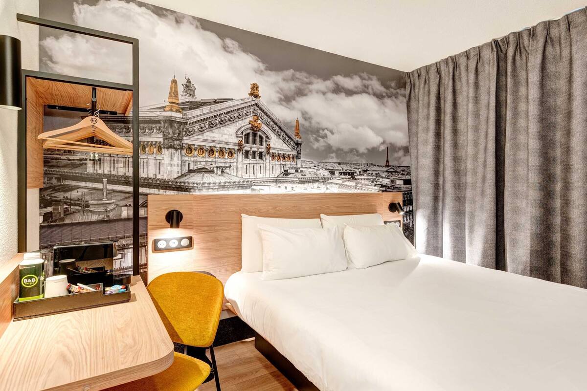 A hotel room with a bed, a desk with a chair, a coffee machine, and a mirror.