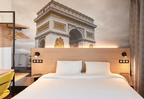 Una habitación de hotel con cama, lámparas de noche, cabecero de madera, escritorio con silla y un mural en la pared del Arco del Triunfo.
