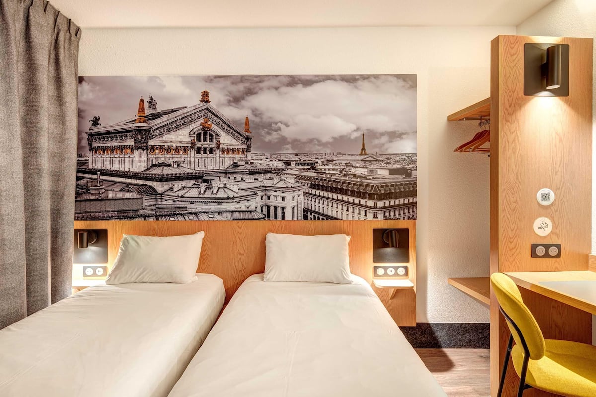 A hotel room with two beds, a desk, a chair, a wall-mounted TV, and a mural of a cityscape.