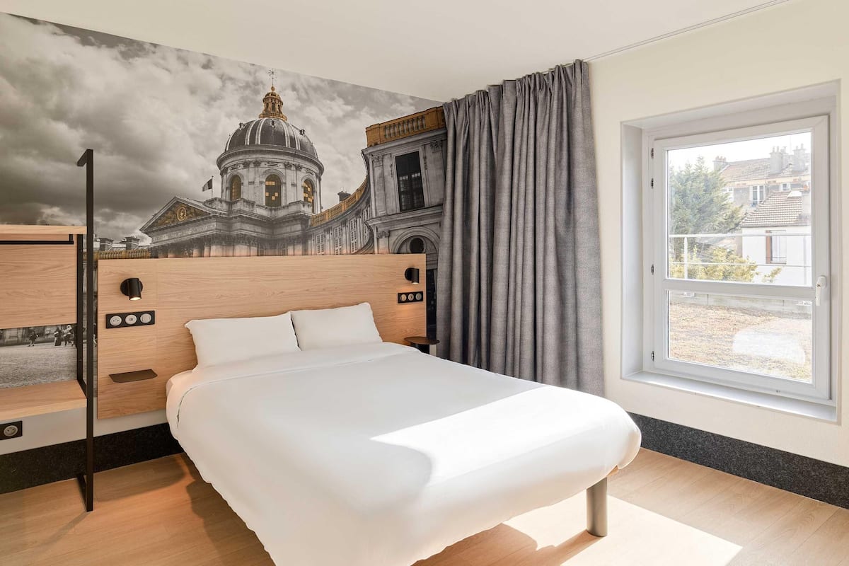 A hotel room with a large bed, a mural of a historical building, a window with a view, and a wooden headboard.