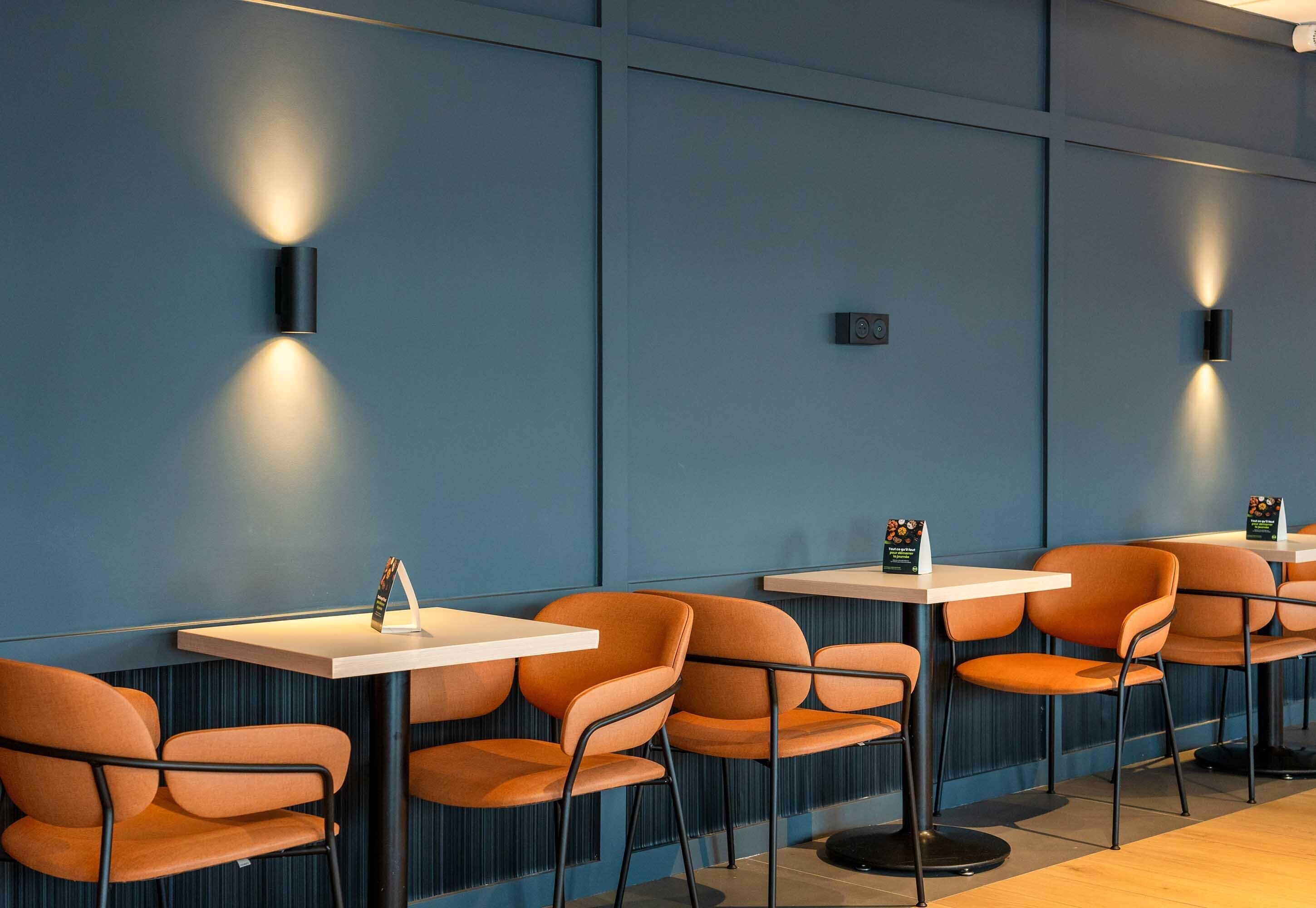 A row of orange chairs and white tables in a modern dining area with blue walls and wall-mounted lights.