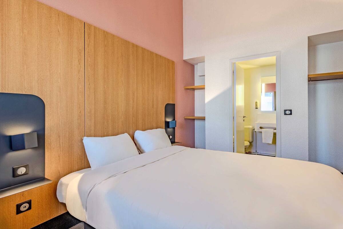 A modern hotel room with a large bed, a wooden headboard, bedside controls, and a bathroom visible through an open door.