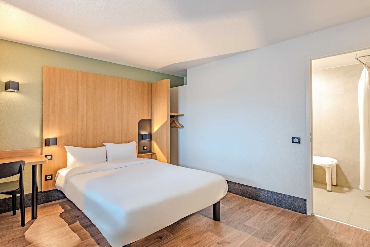 A modern hotel room with a large bed, a wooden headboard, a desk with a chair, and a small table.