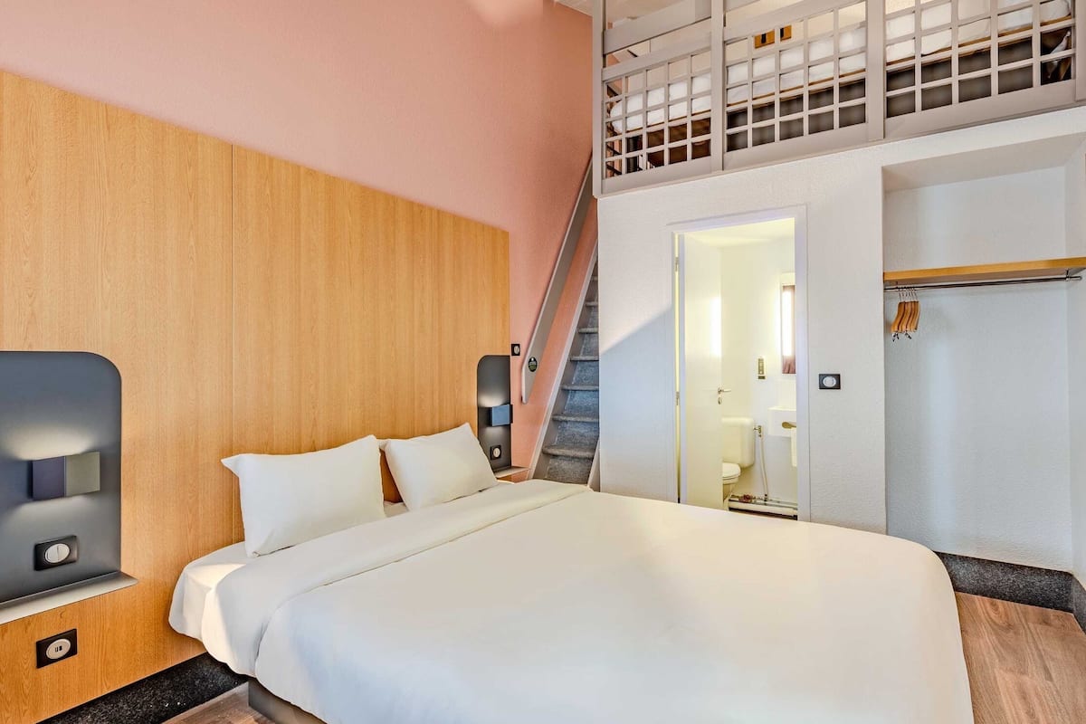 A modern hotel room with a large bed, a wooden headboard, a staircase leading to an upper level, and a bathroom visible through an open door.