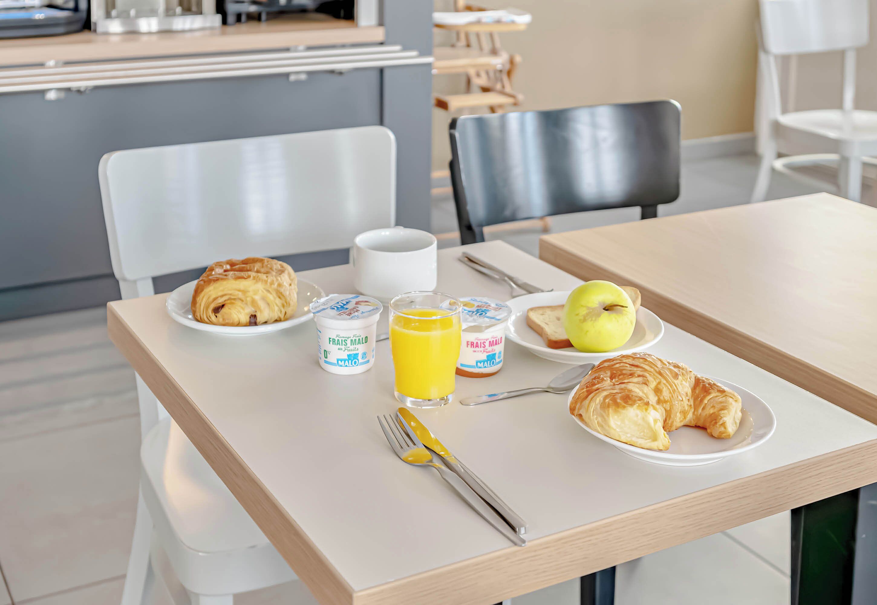 Una mesa de desayuno con croissants, un vaso de zumo de naranja, yogures y un plato con una manzana.