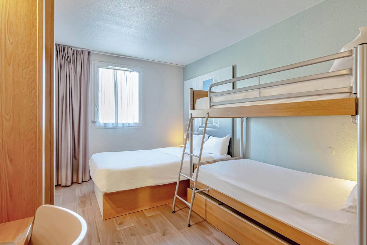 A small, clean room with two bunk beds, a wooden desk, a chair, and a window with curtains.