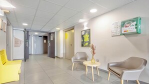 Uma área de recepção moderna com piso ladrilhado, um balcão de atendimento e disposição de assentos, incluindo uma cadeira amarela, um poltrona cinza e uma poltrona marrom.