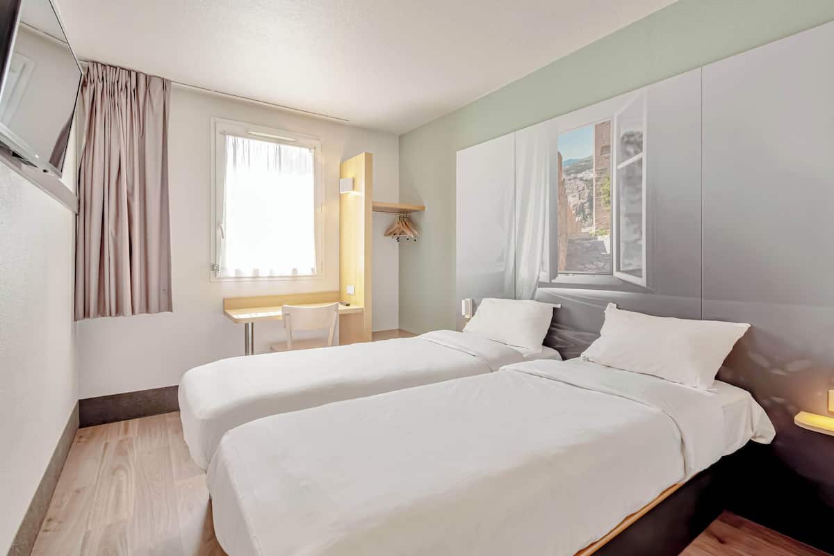 A hotel room with two beds, a desk, a chair, and a window with a view.