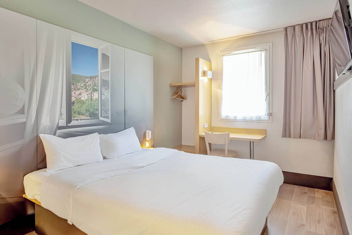 A hotel room with a bed, a desk, a chair, a window with curtains, a coat rack, and a view of a town.