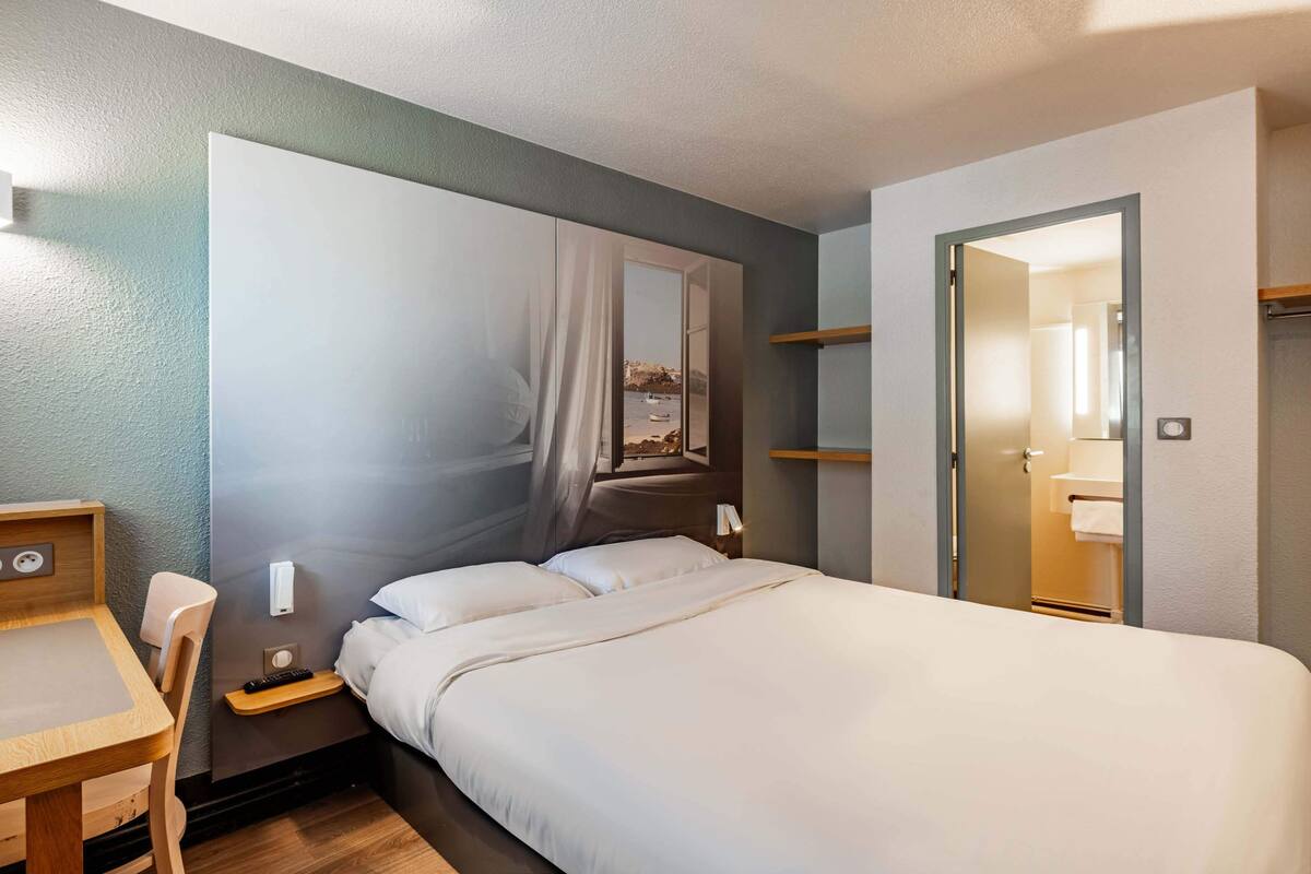 Une chambre d’hôtel comprenant un lit, un bureau, une chaise, une table de chevet, un miroir et une porte donnant sur une salle de bain.