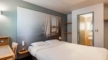 Hotellihuone, jossa on sänky, työpöytä, tuoli, yöpöytä, peili ja ovi, joka johtaa kylpyhuoneeseen.