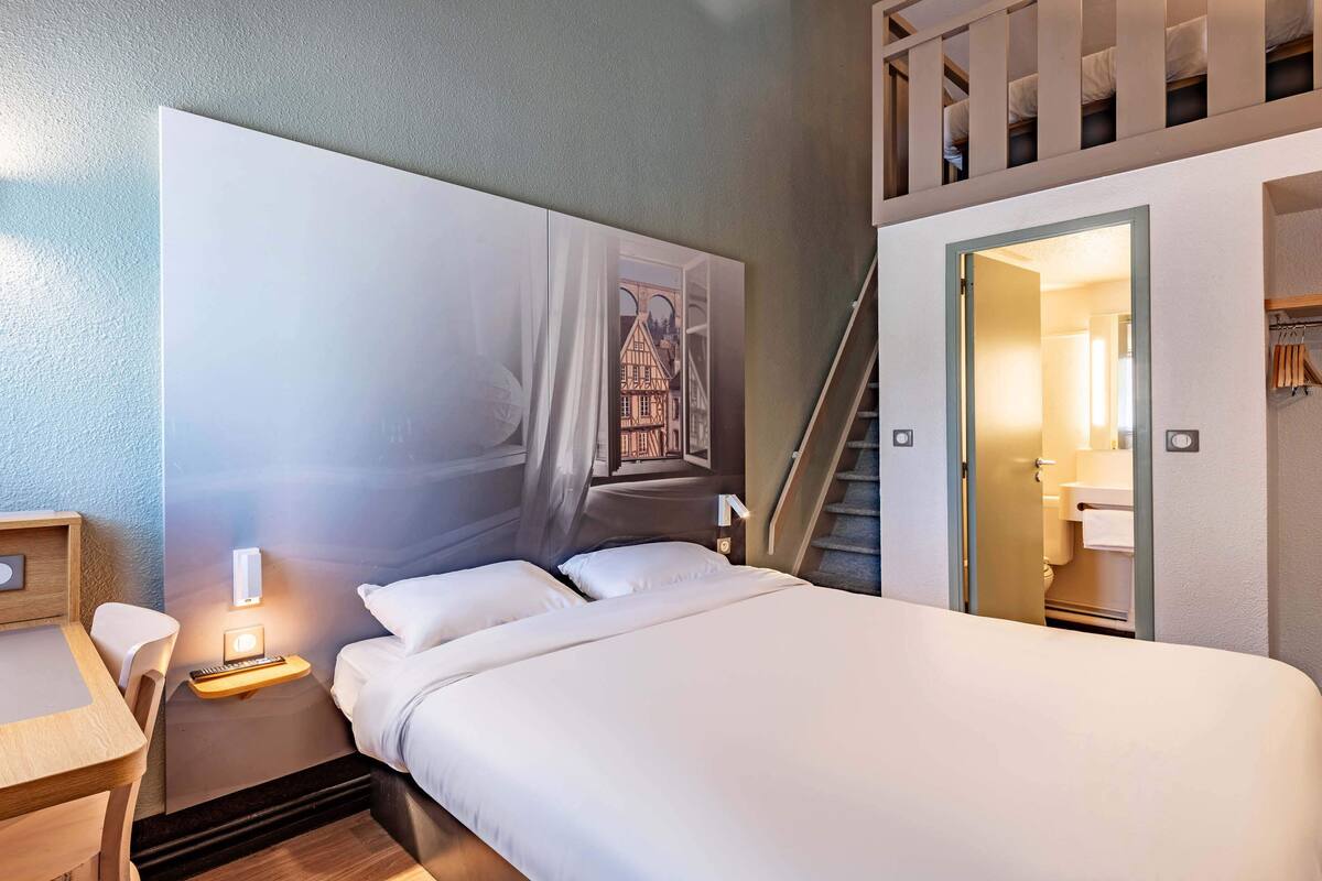 Une chambre d’hôtel avec un grand lit, une table de chevet avec une lampe, une œuvre d’art accrochée au mur, un escalier et une salle de bain visible à travers une porte ouverte.