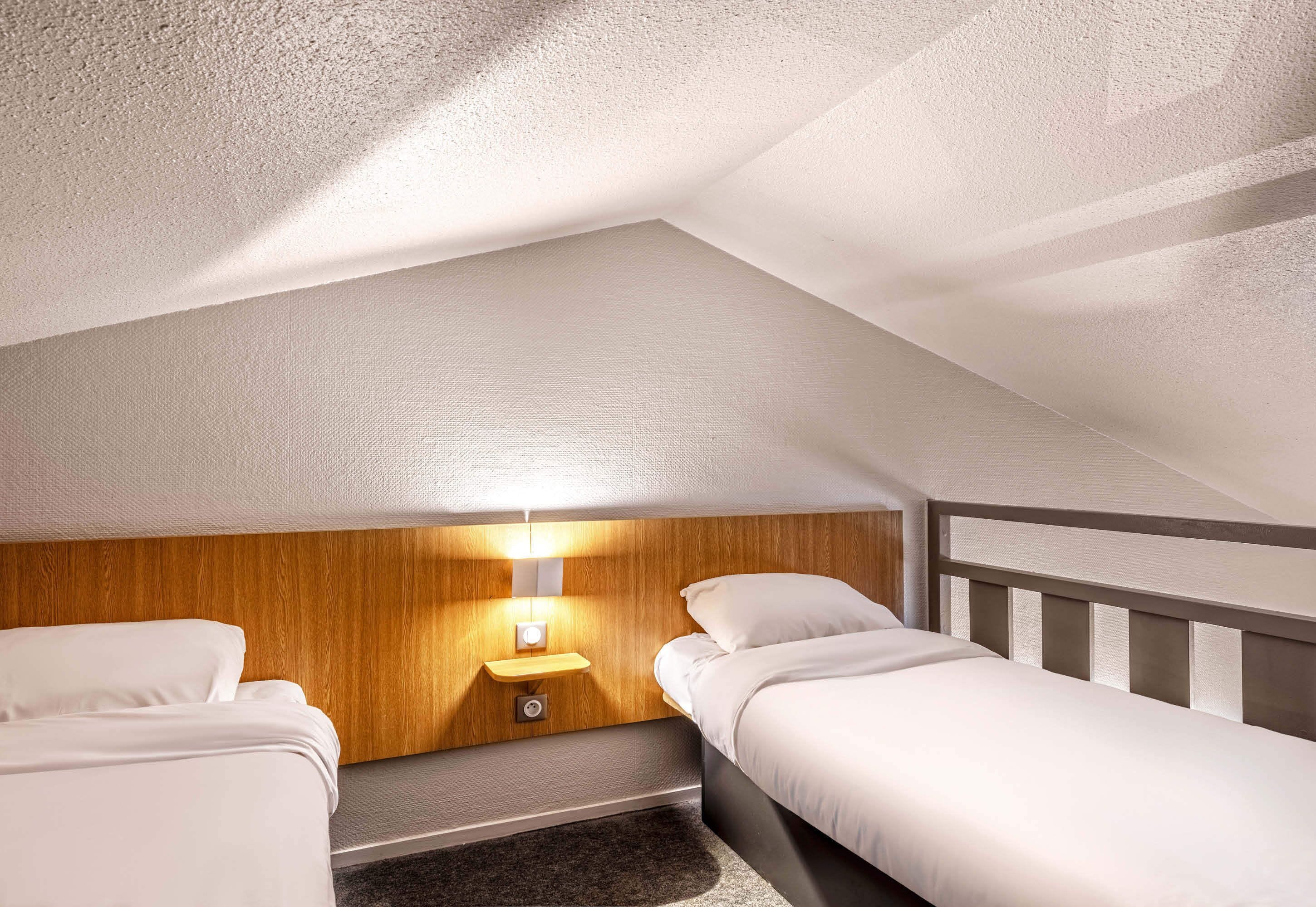 A small, triangular room with two single beds, a wooden headboard, and a nightstand with a lamp.