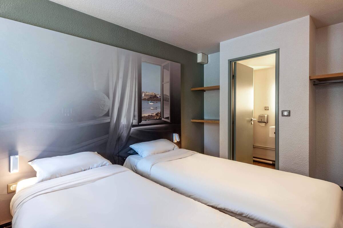 Une chambre d’hôtel avec deux lits, un grand tableau au mur, une porte donnant sur une salle de bain et une étagère avec un miroir.