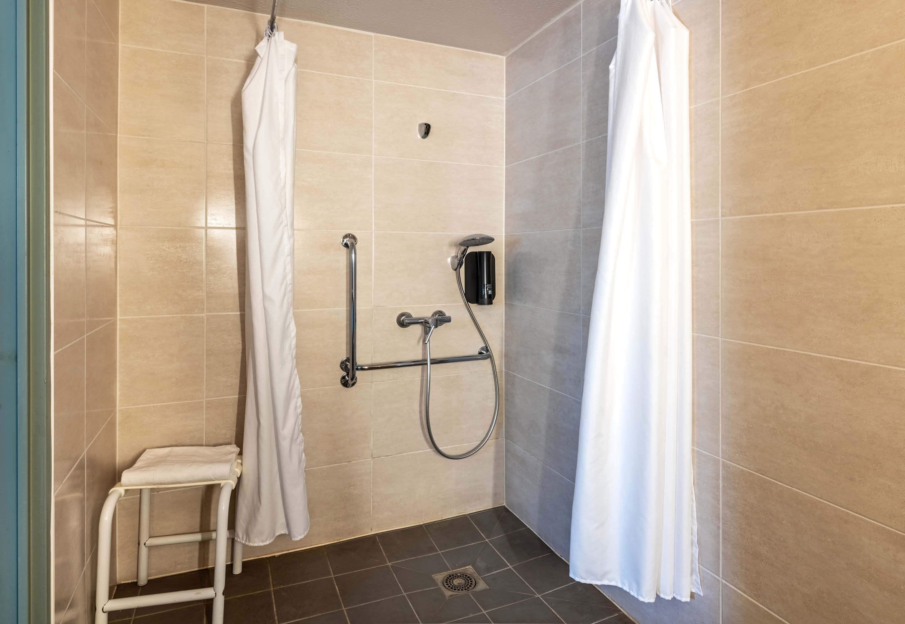 Un área de ducha pequeña con azulejos, cortina blanca, cabezal de ducha manual y un taburete.