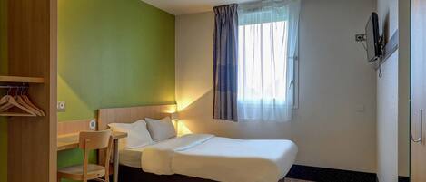 Une petite chambre à coucher, bien rangée, avec un lit, un bureau et une chaise. On y trouve une fenêtre avec des rideaux, un téléviseur fixé au mur et une armoire en bois.