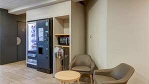Un intérieur moderne avec un distributeur automatique de boissons en libre-service, une poubelle et deux fauteuils en cuir disposés autour d’une petite table en bois.