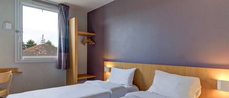 A hotel room with two single beds, a window with curtains, a wooden headboard, a nightstand with a lamp, and a view of buildings outside.