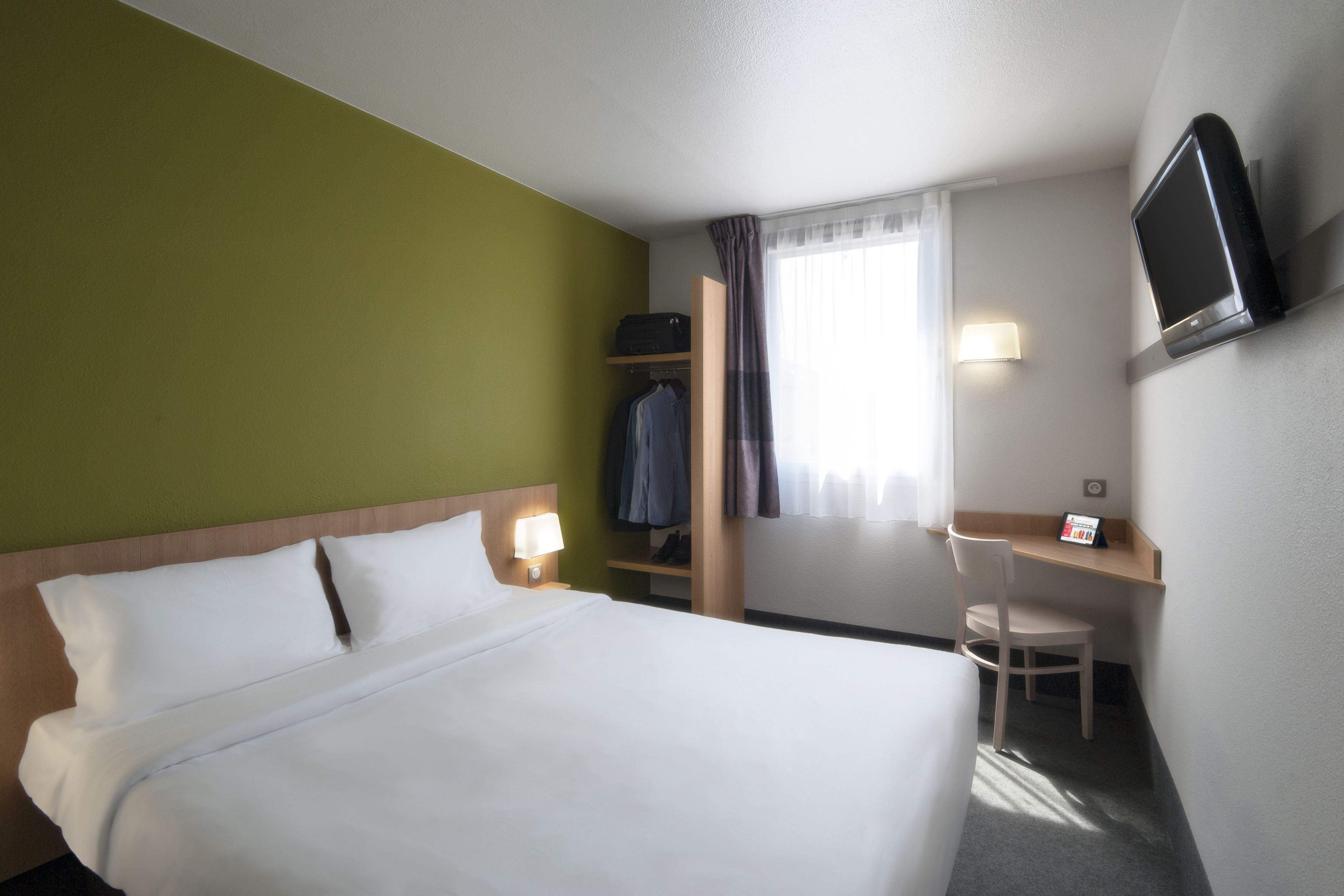 A hotel room with a bed, a desk, a chair, a TV, and a window with curtains.