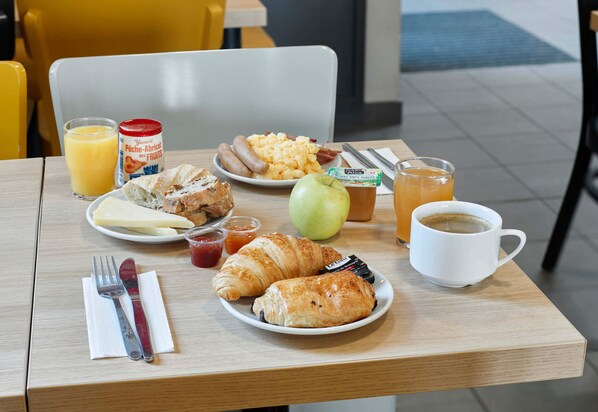 Una mesa de desayuno con cruasanes, salchichas, huevos revueltos, una manzana verde, jugo y una taza de café.