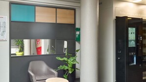 Een moderne lobby met een grijze fauteuil, een klein rond tafeltje, een potplant, een grote zuil en een automaat.