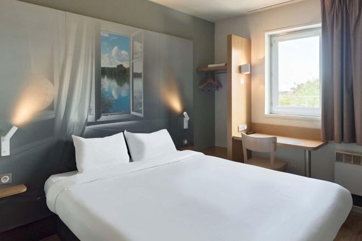 Une chambre d’hôtel avec un lit, un bureau, une chaise, une fenêtre avec des rideaux et un tableau au mur.