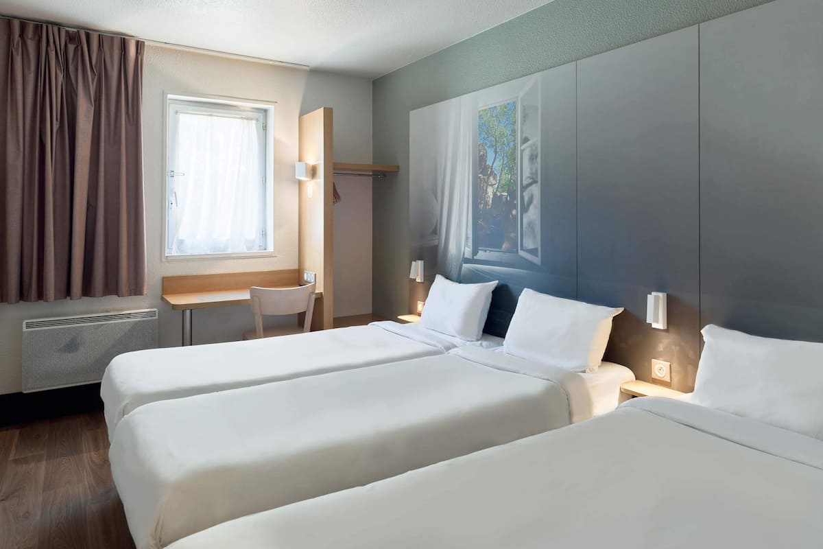 Une chambre d’hôtel avec deux lits, un bureau, une chaise, une fenêtre avec des rideaux et un luminaire fixé au mur.