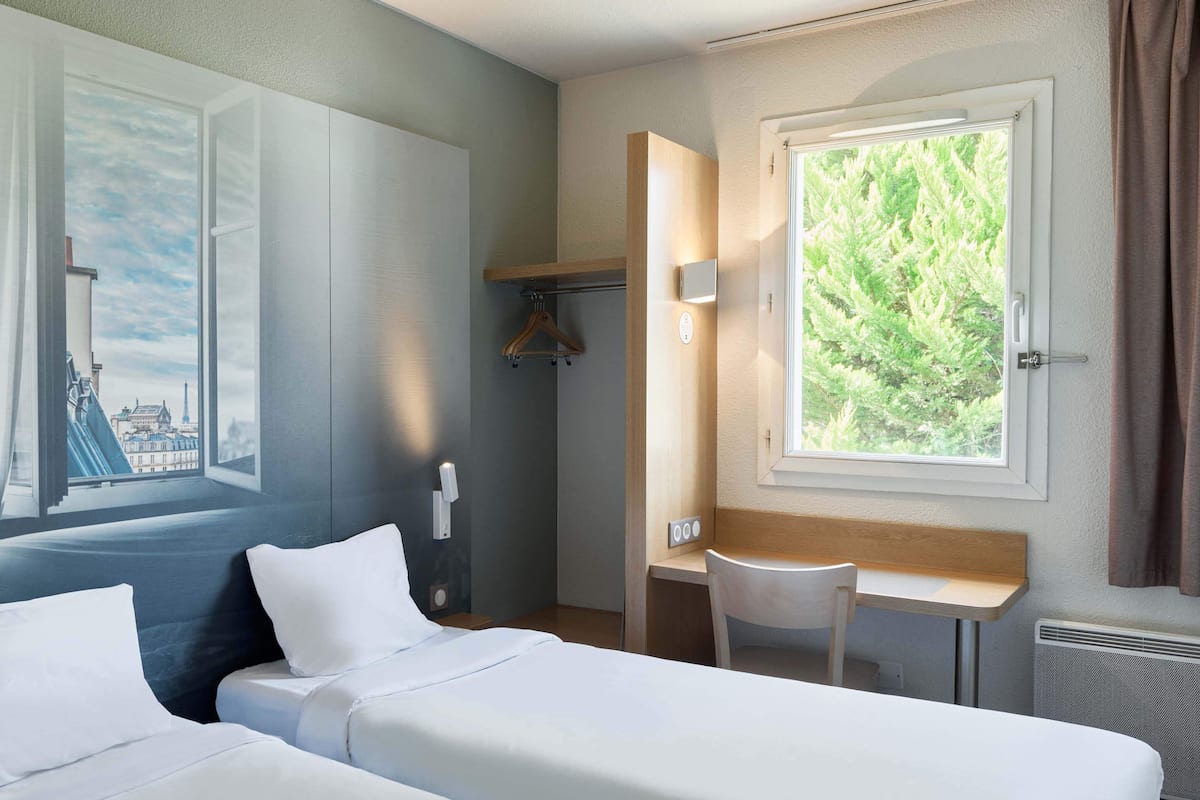 Une chambre d’hôtel avec deux lits, un bureau et une chaise. Il y a une fenêtre donnant sur un paysage verdoyant.