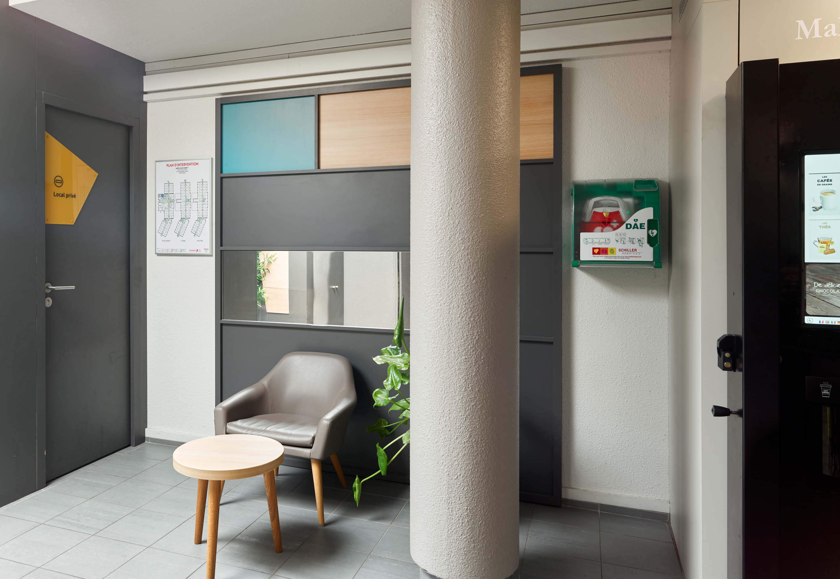 Un moderno vestíbulo de hotel con una puerta gris, una zona de estar con una silla y una mesa redonda pequeña, un pilar grande y un tablón informativo en la pared.