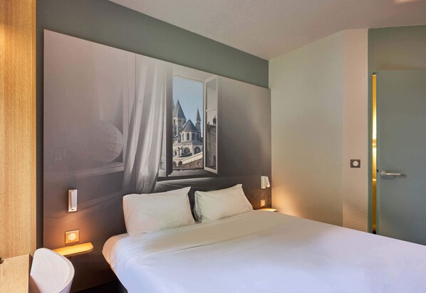 A hotel room with a large bed, two bedside tables with lamps, a view of a building through a window, and a door to the right. - B&B HOTEL Caen Mémorial (Saint-Contest)