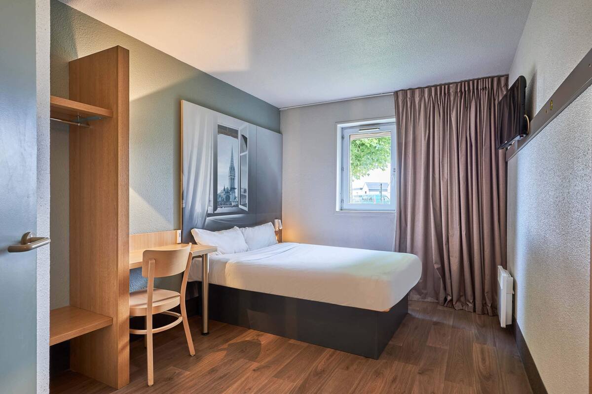 A compact hotel room with a bed, a desk, a chair, and a window with curtains.