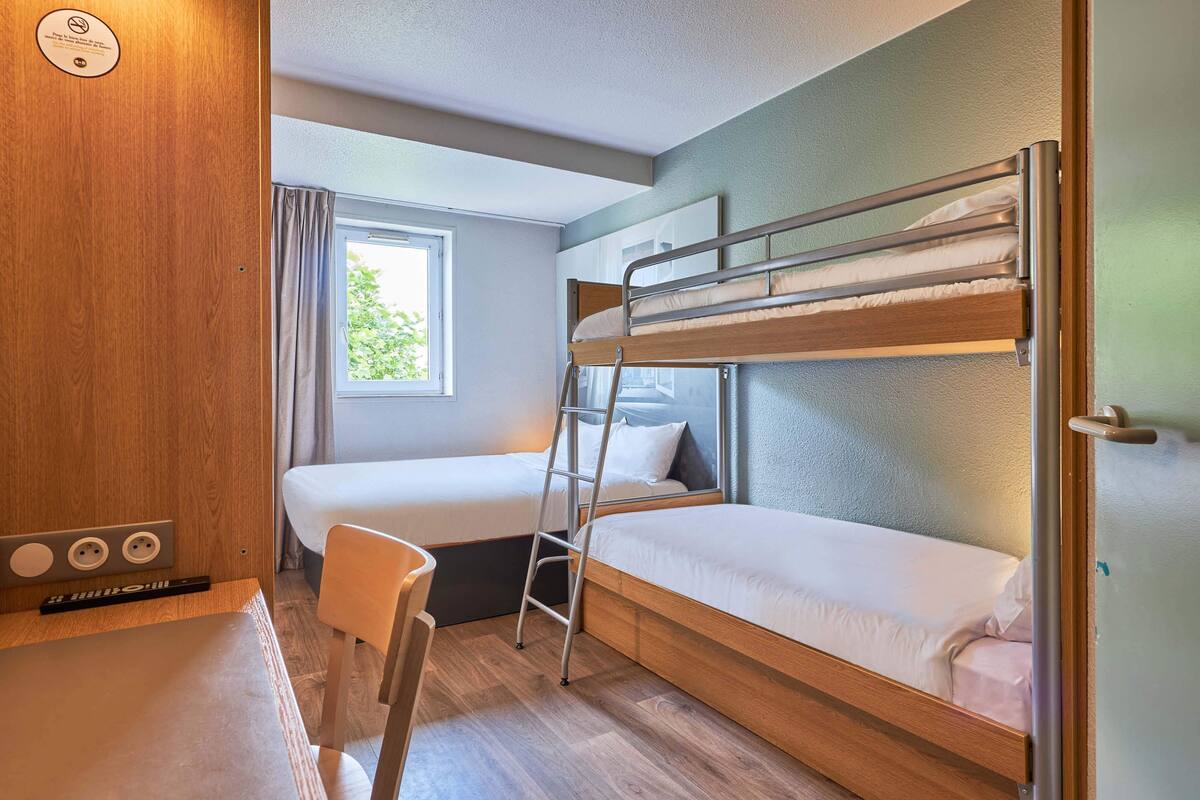 A bunk bed room with a desk and chair, a window with curtains, and a wooden wall.