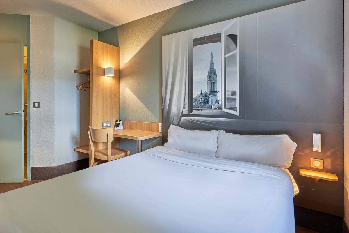 A hotel room with a bed, a desk, a chair, and a view of a building through a window.