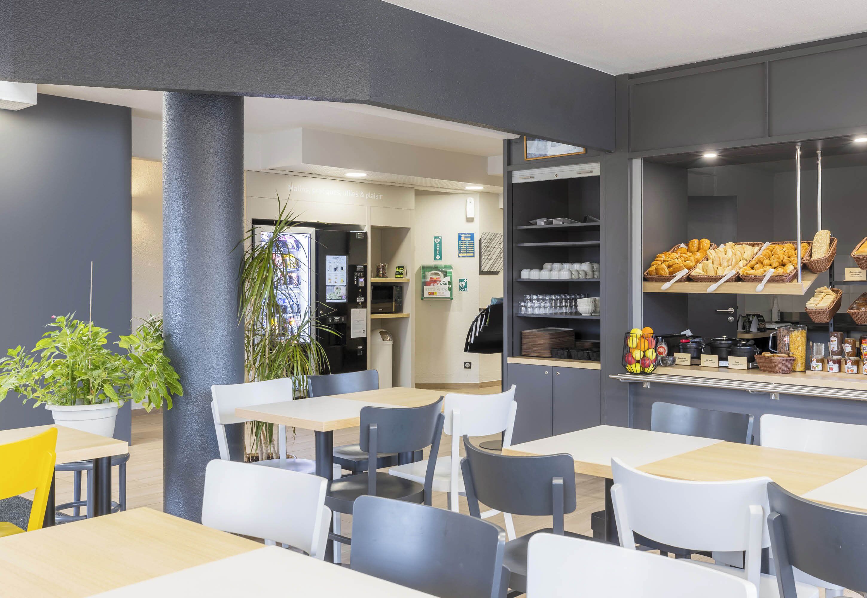 A modern cafe with tables and chairs, a counter with baked goods, and a plant in the corner.