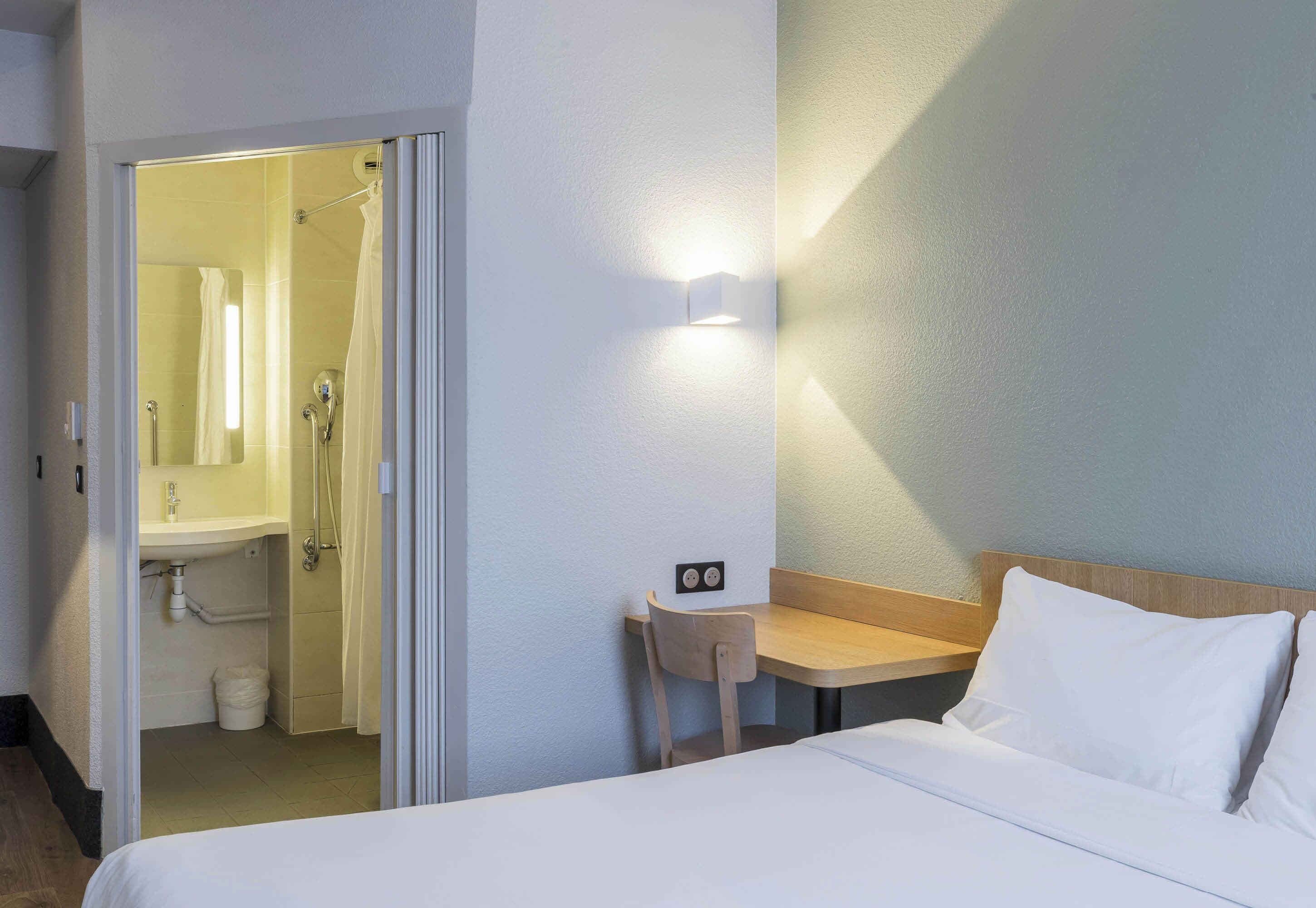 A hotel room with a bed, a small table, a chair, a bathroom with a shower, and a wall-mounted light.