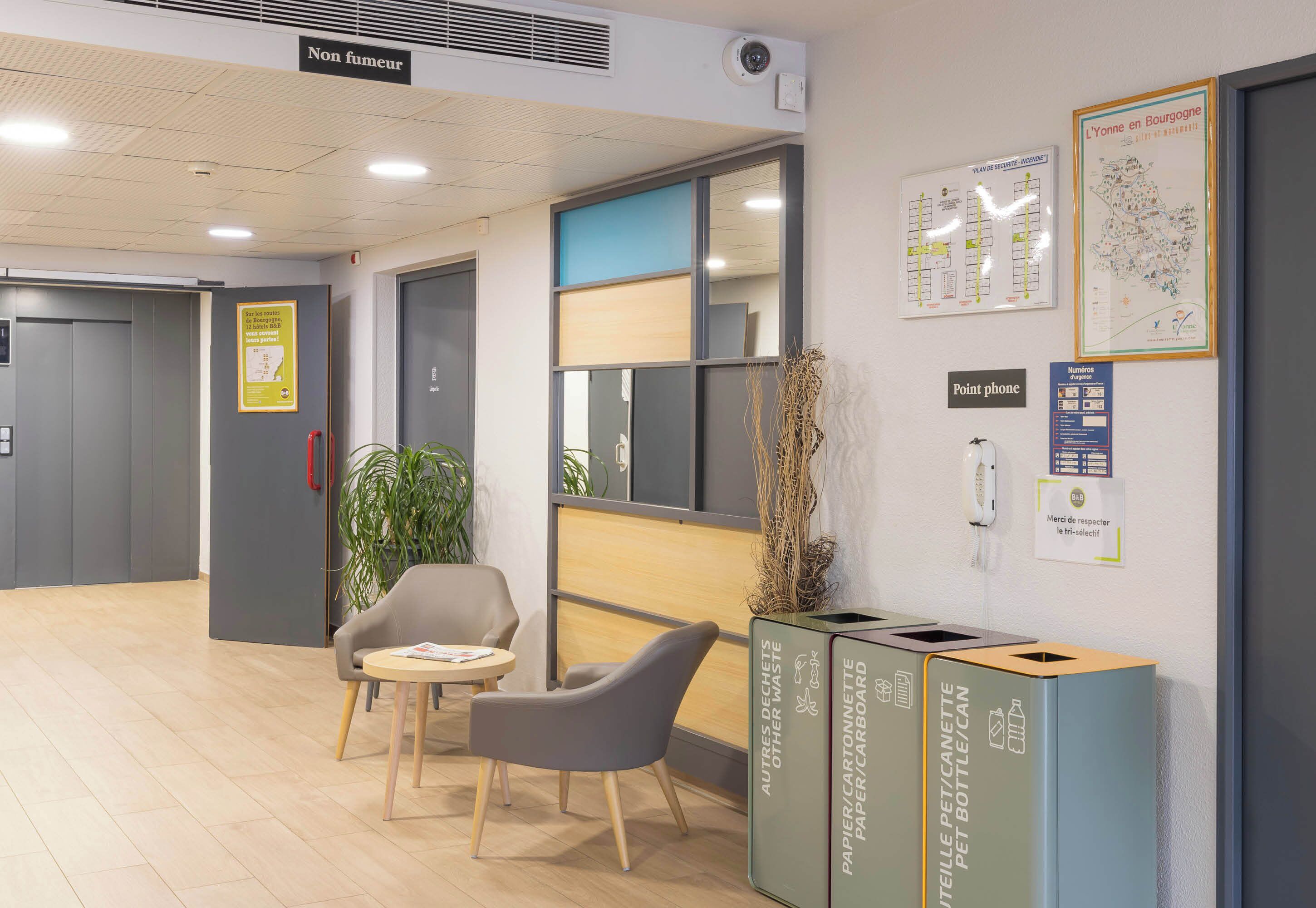 A modern office space with a reception area, including a desk, chairs, a plant, recycling bins, and informational posters on the walls.