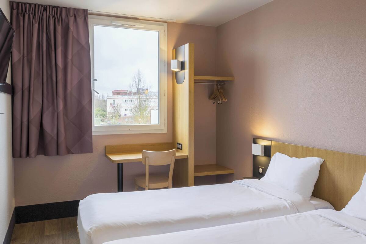 A hotel room with two beds, a desk, a chair, a coat rack, and a window with curtains.