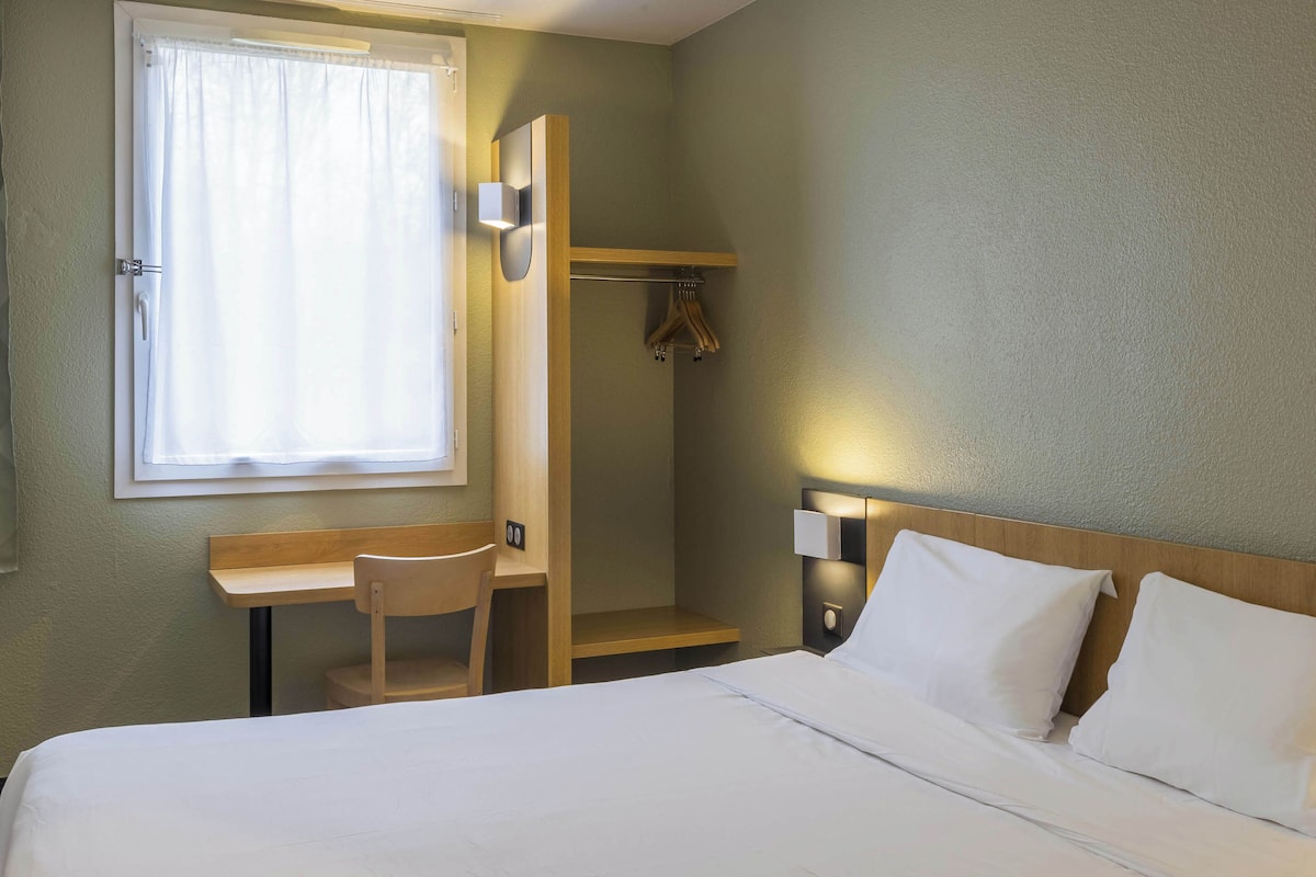 A hotel room with a bed, a desk, a chair, a coat rack, and a window with curtains.