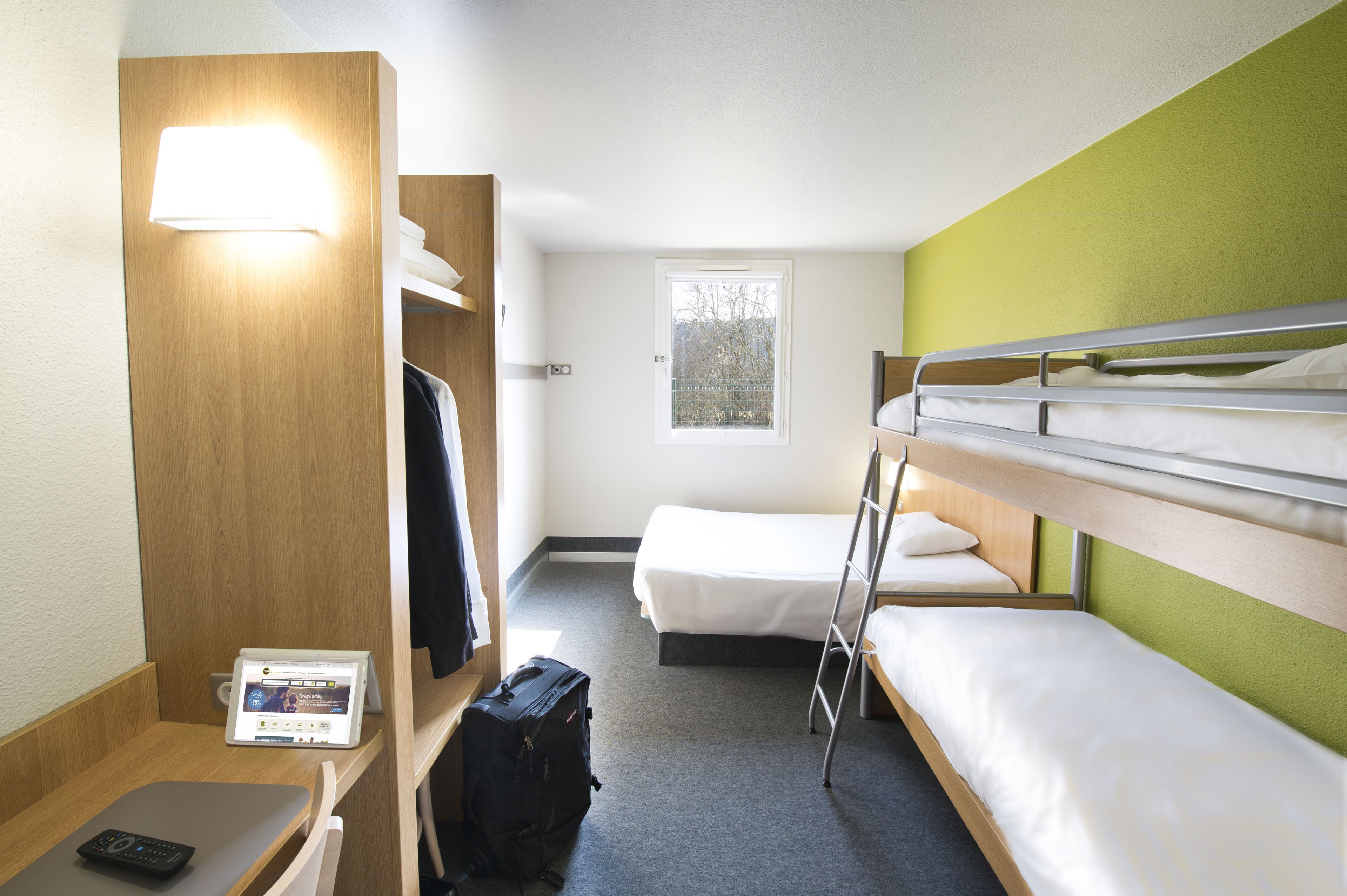 Habitación de hotel con literas, un escritorio con una computadora portátil, una maleta y una ventana con vista a árboles.